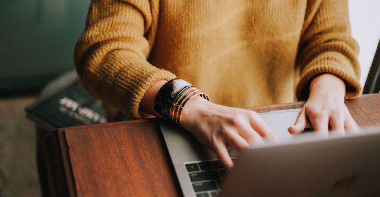 A graduate student in an orange sweater takes classes online with a laptop