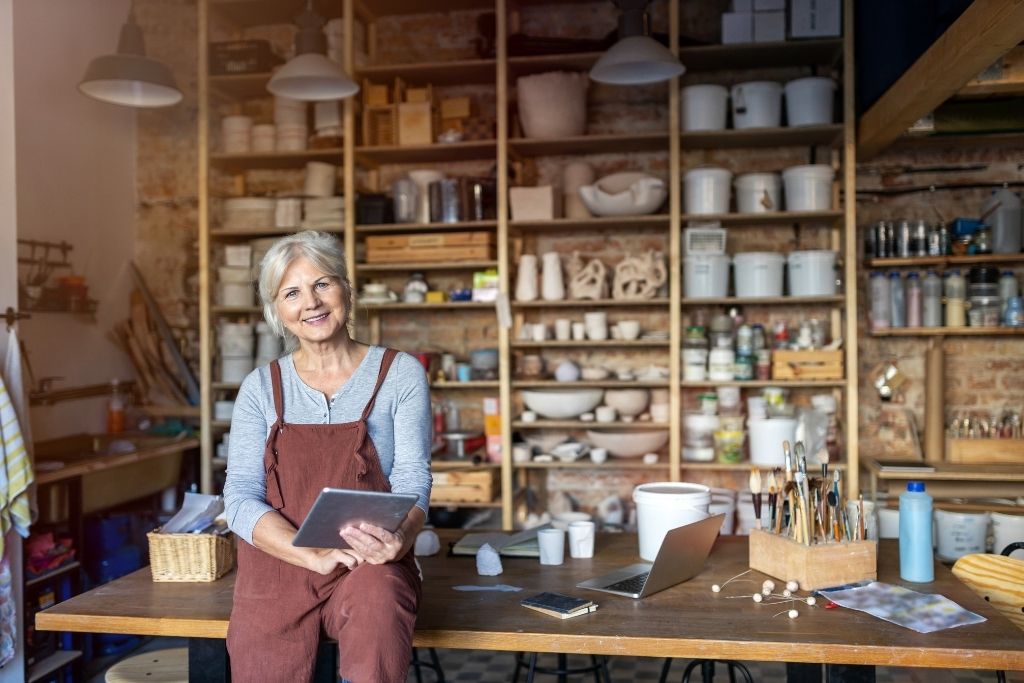 Woman sitting on a wood table with a tablet in her hand and laptop and crafts on the table.