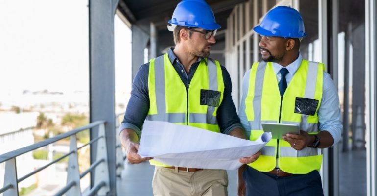 Two male construction managers discuss blueprints on a job site.