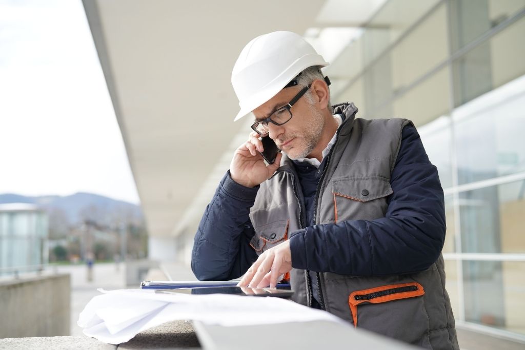 An older male construction manager overlooks plans while talking on a cell phone at the job site.