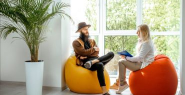 A female health coach sits on a bean bag chair while talking with a male client.