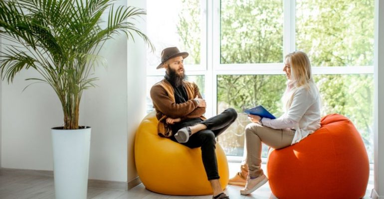 A female health coach sits on a bean bag chair while talking with a male client.