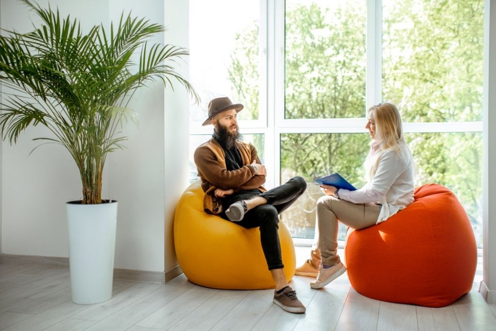 A female health coach sits on a bean bag chair while talking with a male client.