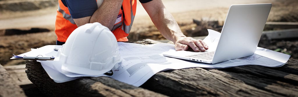 A surveying manager looking at a computer outside with plans under it.