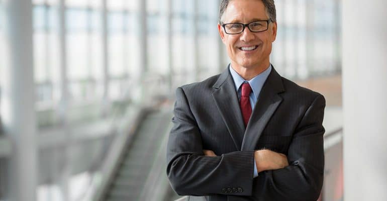 Man wearing a suit standing inside an airport with an escalator behind him.