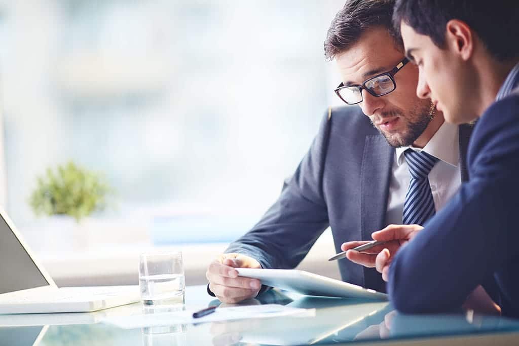 Two businessmen talk while looking over an iPad with a laptop in the background.