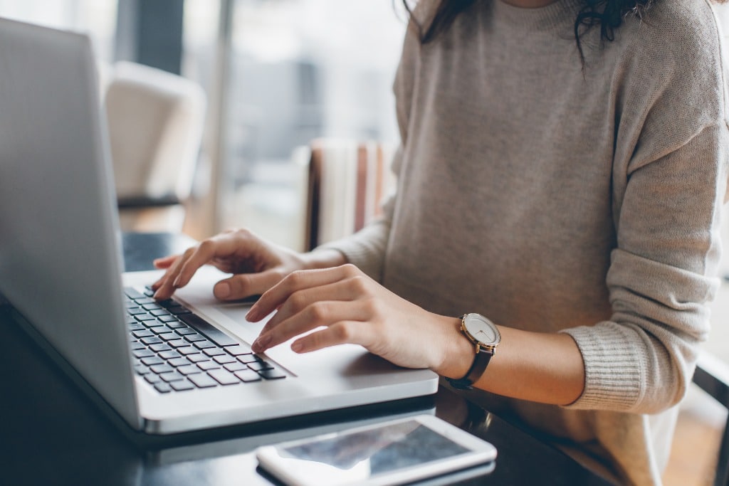 A college student types on her laptop with her phone on the desk.