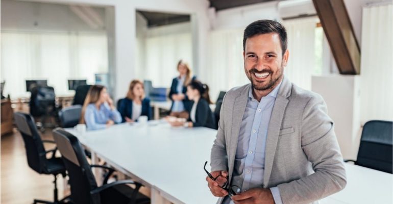 A smiling young male entrepreneur leans on a white conference table during a meeting.