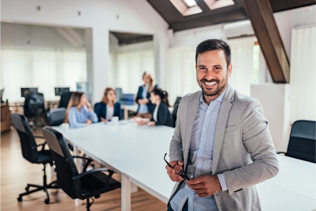 A smiling young male entrepreneur leans on a white conference table during a meeting.