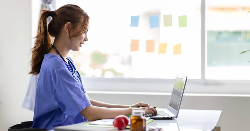 Health sciences student in scrubs studying on a laptop at a desk