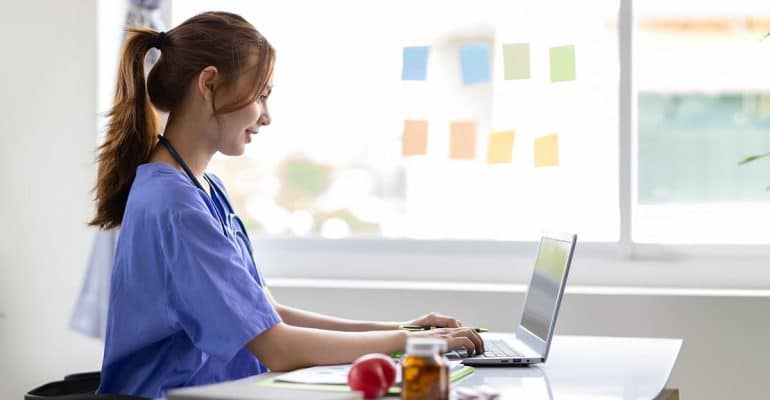 Health sciences student in scrubs studying on a laptop at a desk