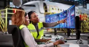 Two aviation workers in hi-vis vests reviewing aircraft data on curved monitors in a hangar