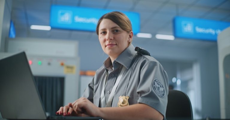 Airport security officer in uniform with a badge working at a laptop beside a checkpoint scanner