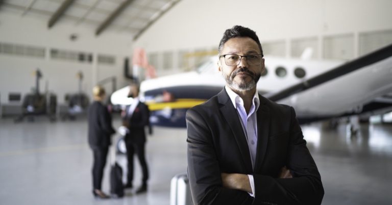 Confident aviation professional in a suit standing in a hangar with a private jet and crew behind him