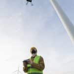 Technician in hard hat and safety vest flying an inspection drone beside a wind turbine at a wind farm