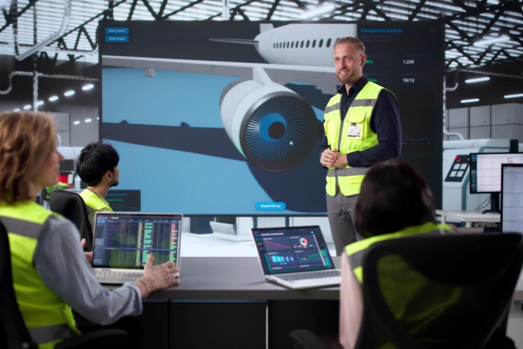 Instructor in a safety vest leading an aviation team meeting with an aircraft engine on a large screen